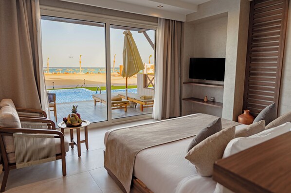 A hotel room with a large bed, a TV, a small table with fruit, and a view of the beach and pool.