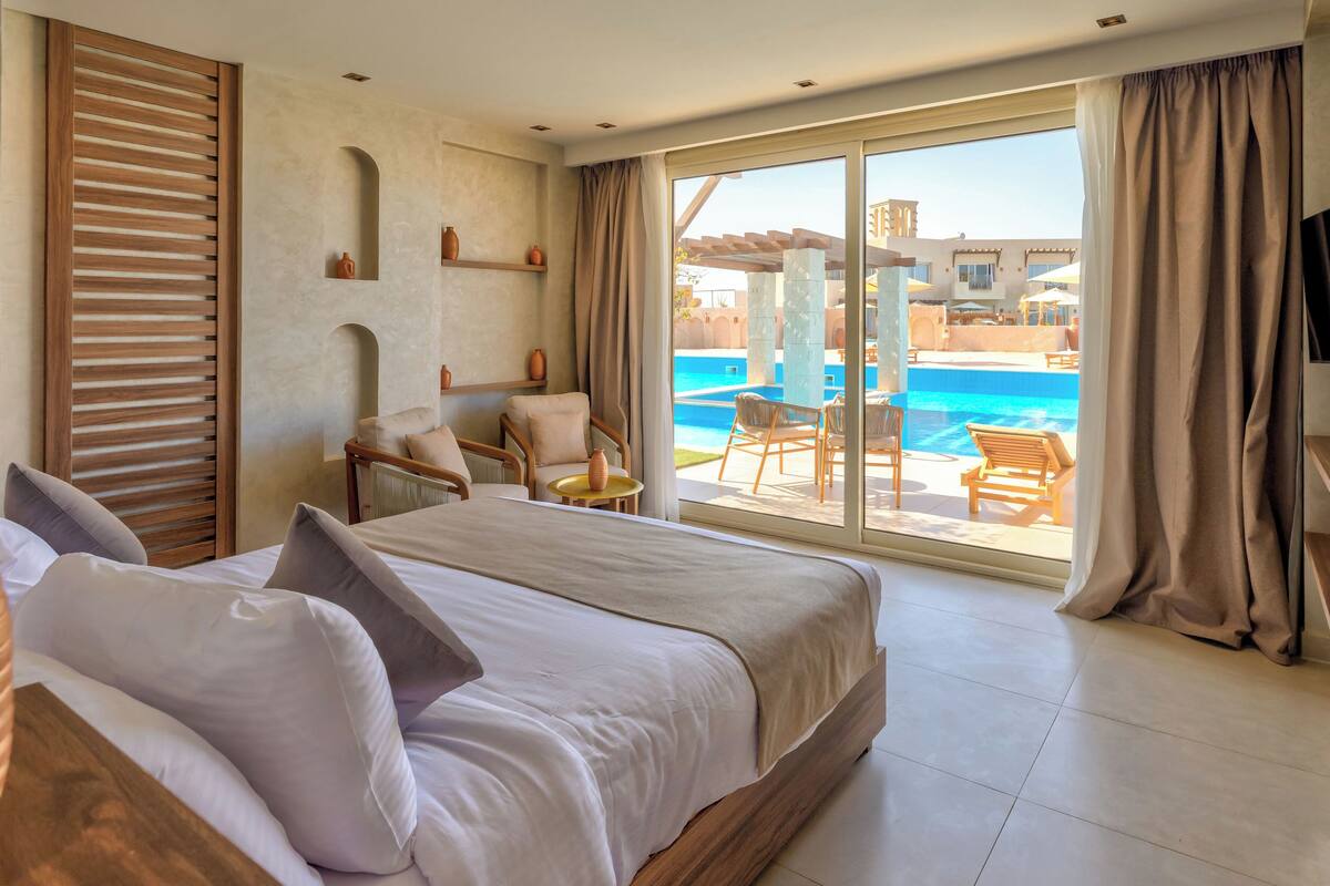 A hotel room with a large bed, a seating area with chairs and a table, and a view of a swimming pool.