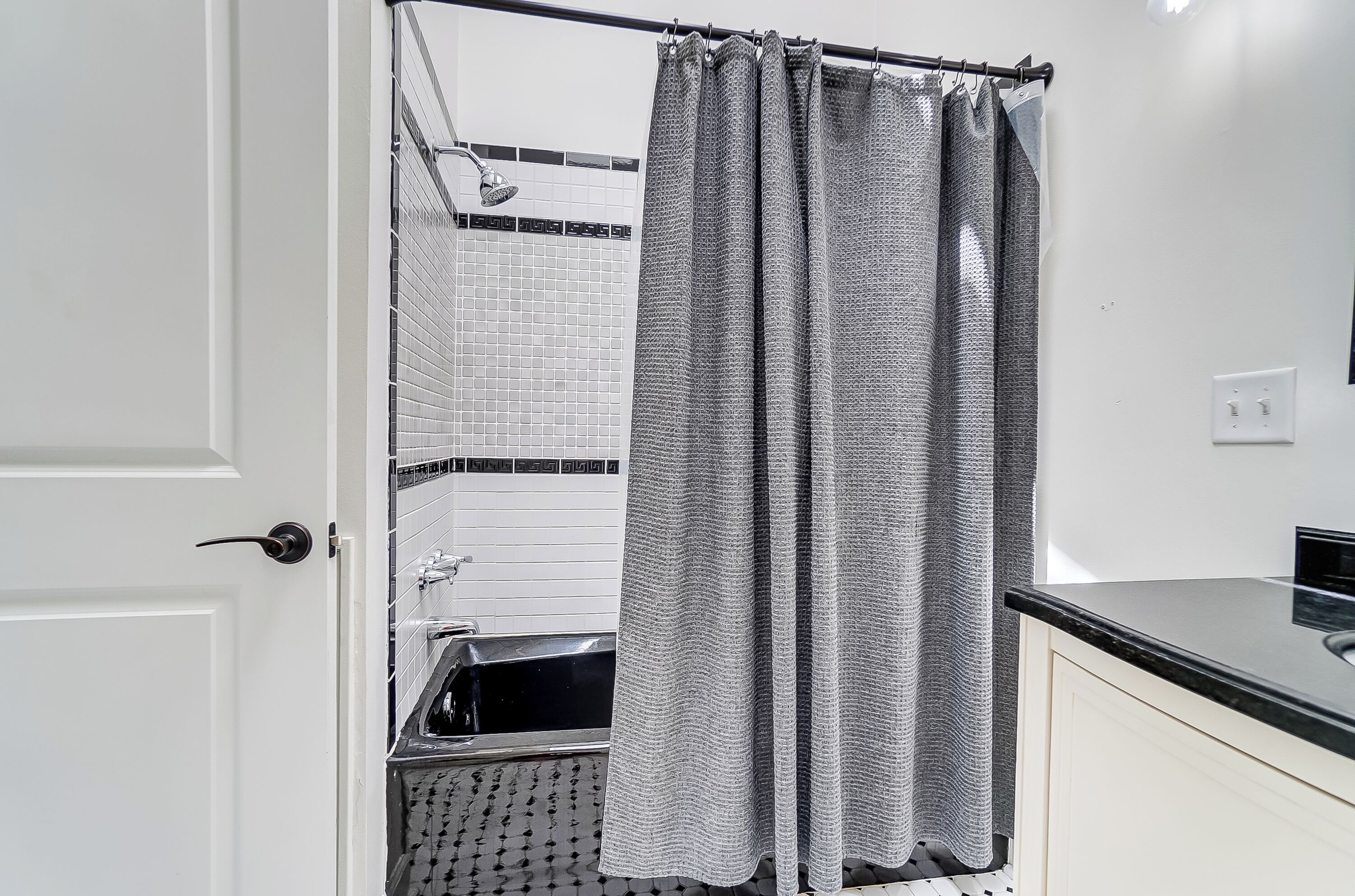 Un baño con ducha, paredes de azulejos blancos, encimera negra y una puerta blanca.