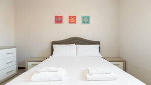 Una cama bien hecha con sábanas y toallas blancas, un cabecero de madera y una mesita de noche con un cajón. Tres carteles enmarcados están colgados en la pared sobre la cama.
