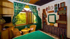 Quarto com cama, duas cadeiras de vime, uma mesinha, televisão e janela com cortinas verdes.