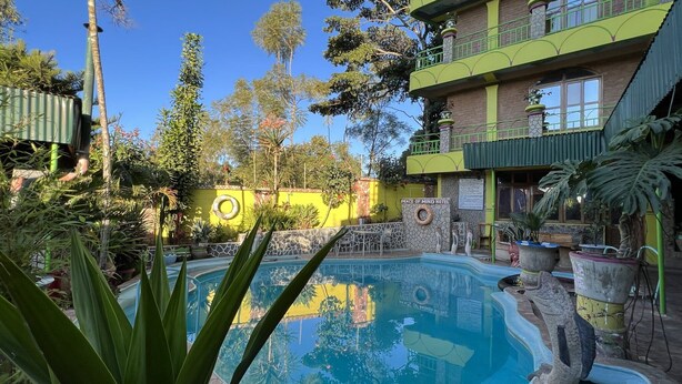 Uma área de piscina com borda de pedra, rodeada por plantas em vasos e um edifício com fachada amarela.