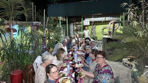 Um grupo de pessoas jantando ao ar livre em uma mesa comprida, com diversos pratos e bebidas.
