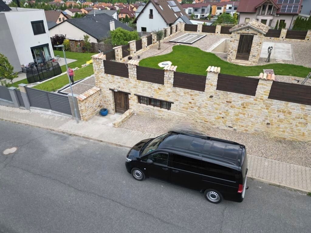 A black van parked on a paved road in a residential area with stone walls and a grassy lawn.
