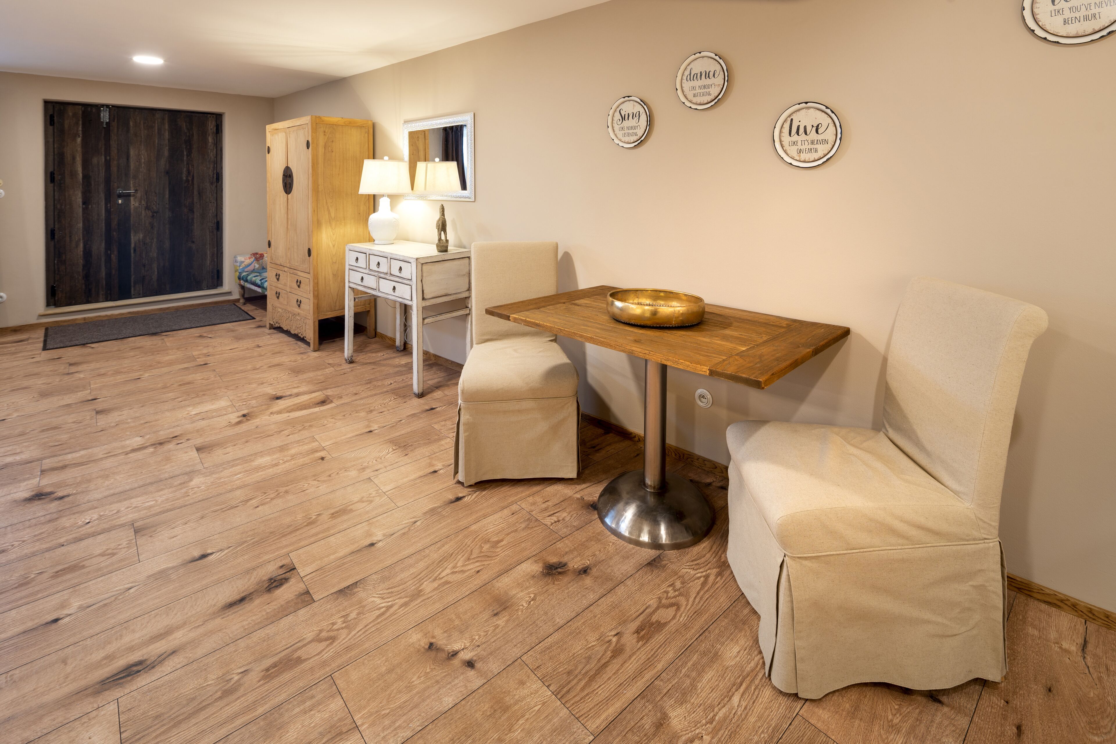 A room with a wooden floor, a dining table with a bowl, two cushioned chairs, a white dresser, a lamp, and a wardrobe.