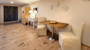 A room with a wooden floor, a dining table with a bowl, two cushioned chairs, a white dresser, a lamp, and a wardrobe.