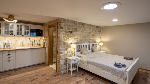A bedroom with a stone wall, a wooden floor, a bed with a white headboard, bedside tables with lamps, and a kitchen area with cabinets and a microwave.