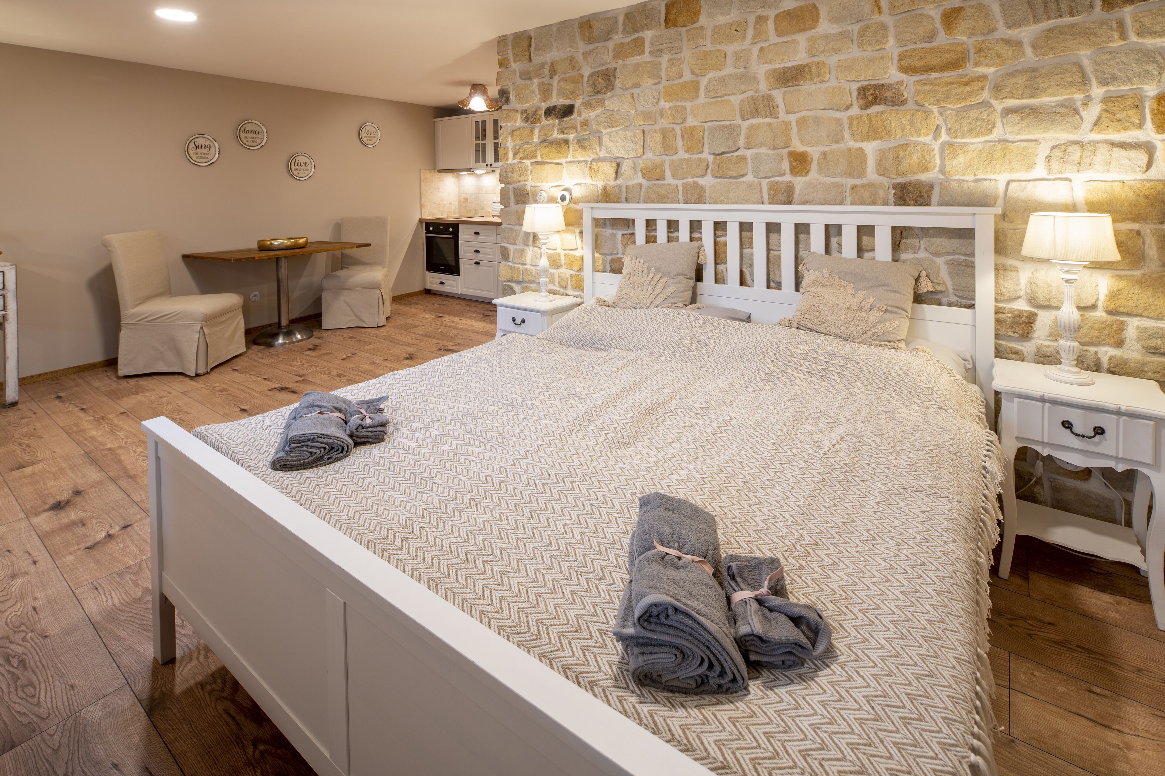 A bedroom with a stone wall, a bed with a herringbone-patterned bedspread, two bedside tables with lamps, a small desk, and a chair.
