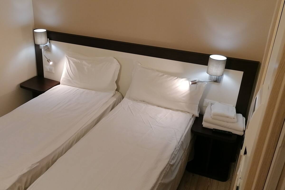 A double bed with white linens, two bedside lamps, and a small table with a folded towel.