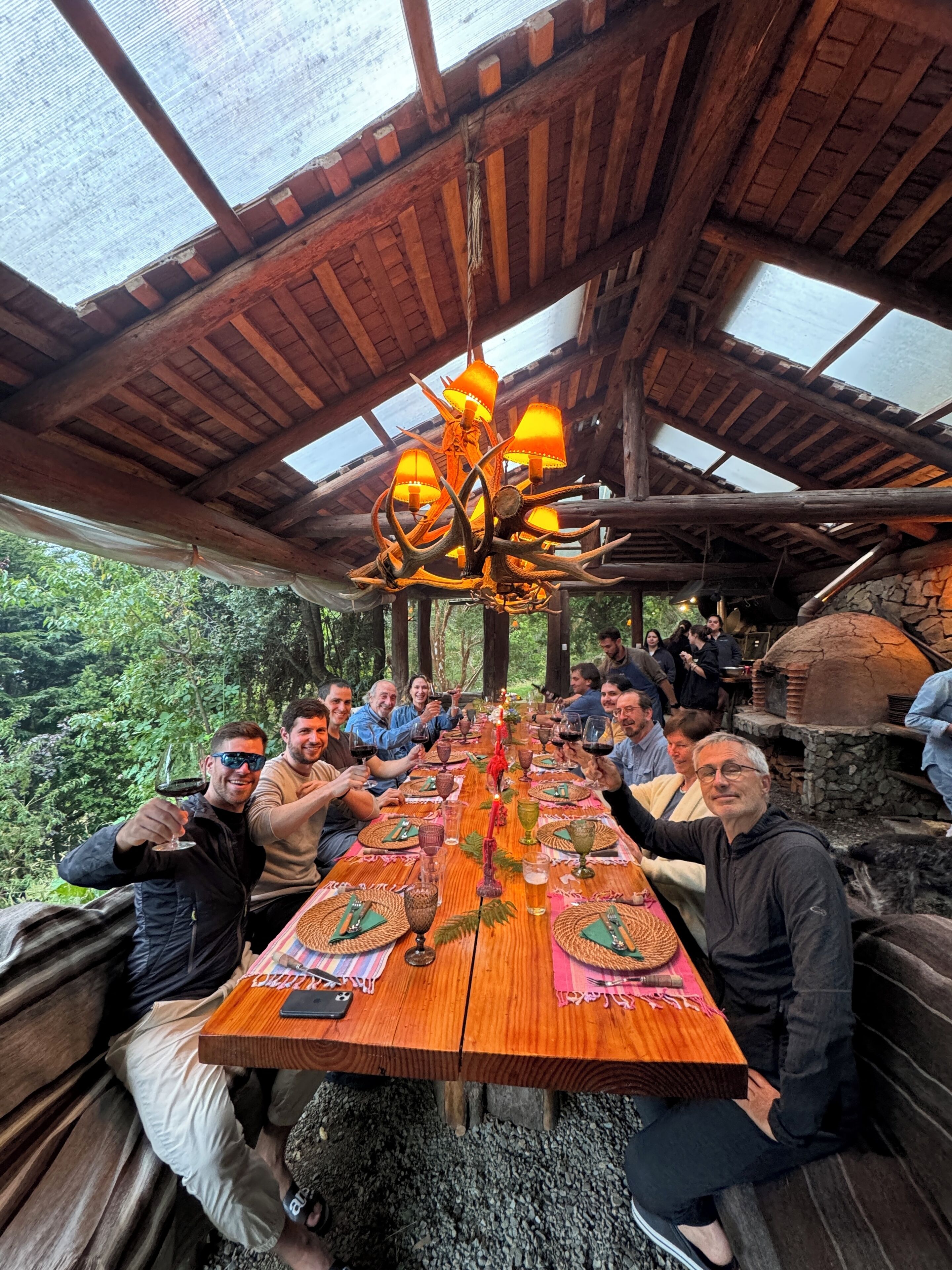 A group of people seated at a long wooden table under a wooden ceiling with a chandelier, surrounded by greenery.