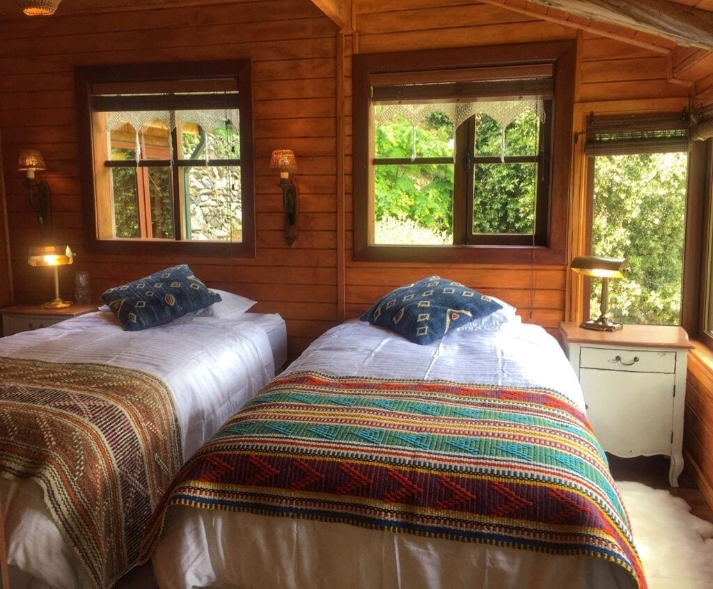 Une chambre dans un chalet en bois, avec deux lits, chacun agrémenté de draps à motifs, deux fenêtres avec des rideaux et une table de chevet avec une lampe.