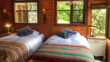 Une chambre dans un chalet en bois, avec deux lits, chacun agrémenté de draps à motifs, deux fenêtres avec des rideaux et une table de chevet avec une lampe.