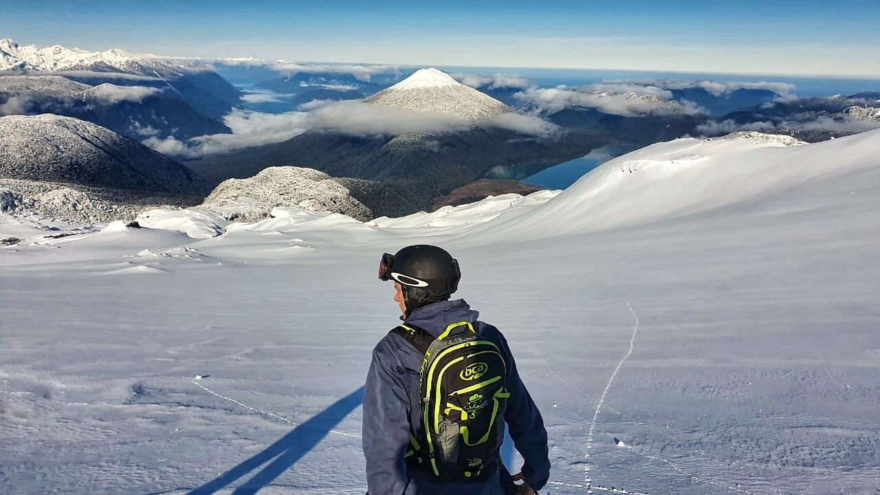 Una persona está de pie en una montaña nevada, con una vista despejada de montañas y nubes distantes.