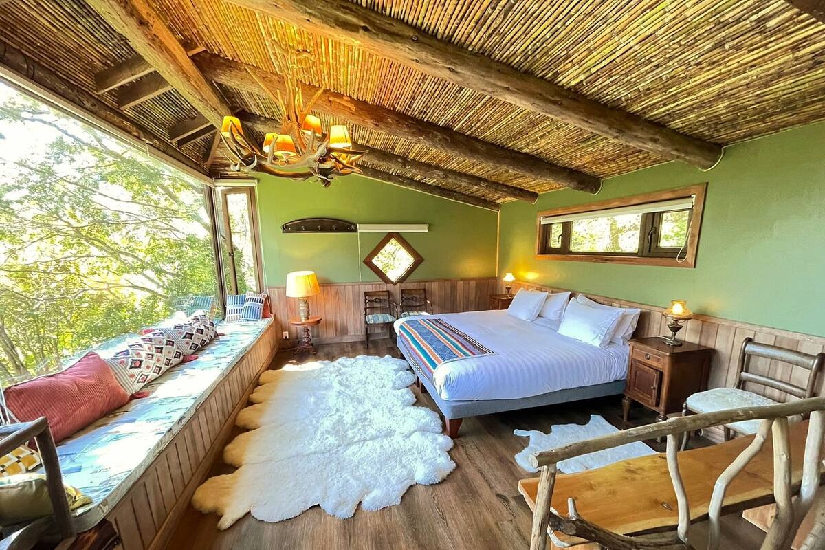 Quarto com cama, um assento junto à janela, piso de madeira, teto de palha e um candelabro.