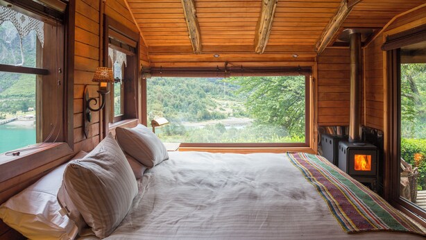 Een houten kamer met een bed, een raam met uitzicht op een bos en een houtkachel.