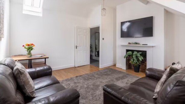 A living room with two leather sofas, a grey rug, a flat-screen TV above a fireplace, and a door leading to another room. - Castle Garden View - Two Steps from Central Rochester High Street (Rochester)