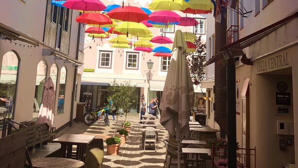 A courtyard with colorful umbrellas hanging overhead, tables and chairs, and a building with a sign reading "CELLA CENTRAL". - Apartment Zachary (Kaprun)