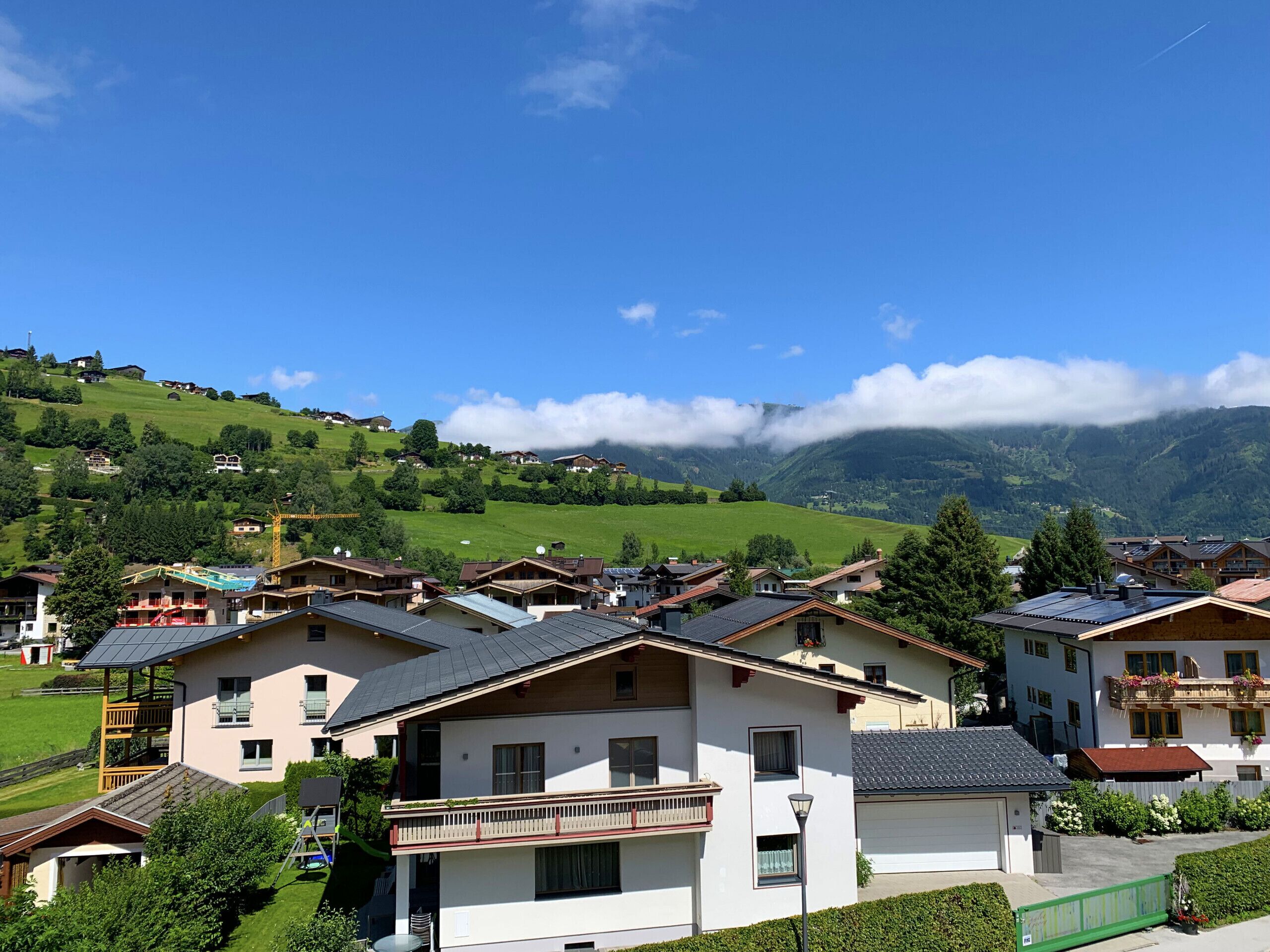 A residential area with houses featuring balconies and solar panels, set against a backdrop of green hills and a mountain range.