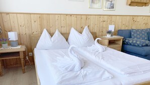 A neatly made bed with white linens and pillows, a wooden headboard, a bedside table with a lamp and a small plant, and a blue sofa with floral cushions in the background.