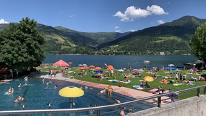 A swimming pool with people, a grassy area with umbrellas, and a lake with mountains in the background.