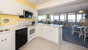 A modern kitchen with white cabinets, a black oven, and a microwave. There's a dining area with a table and chairs, and large windows offering a view. - Beautiful sunset and ocean view from 2 Bedroom Condo with Free WiFi and an Outdoor Pool Located in Midtown Only Steps to Beach! (Ocean City)
