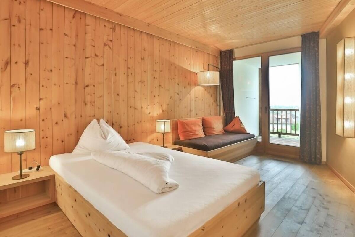 A wooden-paneled room with a bed, a bench, and a window with a view.