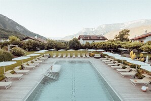 Area piscina con lettini e ombrelloni, circondata da edifici e montagne.