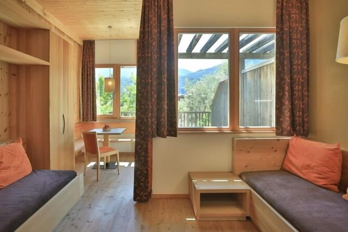 A cozy room with wooden walls and a wooden ceiling, a small table with chairs, a bench with cushions, and a window with curtains.