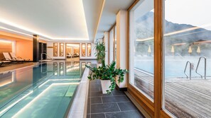 Una piscina interna con pareti in vetro trasparente, un solarium in legno esterno e vista sulle montagne.