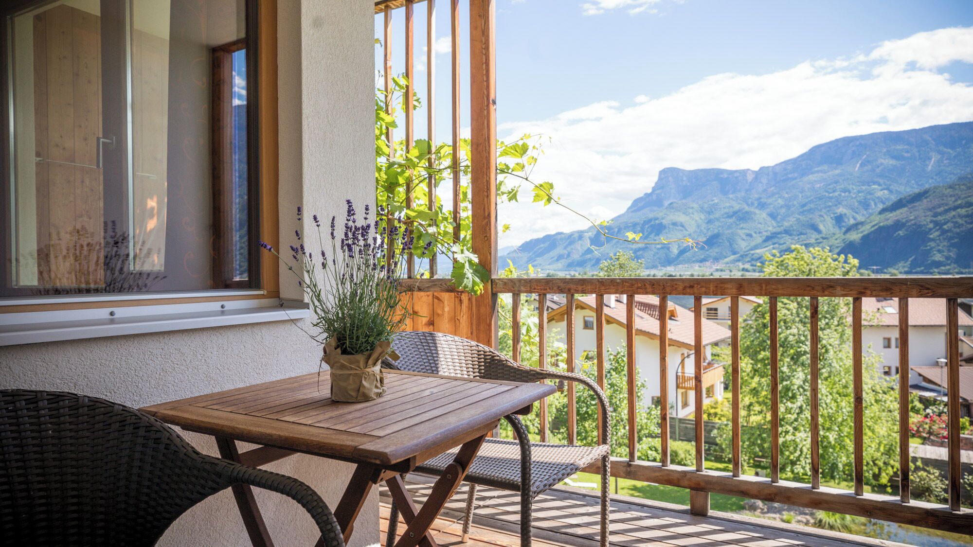 A balcony with a wooden table and chairs, a potted plant, and a view of mountains.