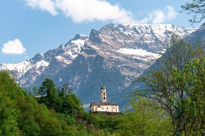 Uma igreja de montanha com um campanário, rodeada por picos nevados e vegetação exuberante.