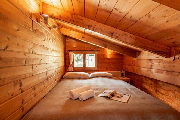 Quarto de cabana de madeira com cama, almofadas, janela e material de leitura.