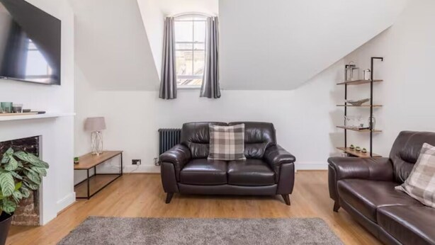 A modern living room with a brown leather sofa, a grey rug, a fireplace, a TV mounted on the wall, and a window with curtains. - Castle Garden View - Two Steps Away from Central Rochester High Street (Rochester)