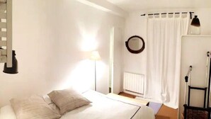 Un dormitorio con cama, lámpara, espejo, radiador y ventana con cortinas.