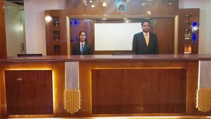 A reception desk with two individuals standing behind it, a wooden panel design, and a blue-lit backdrop. - Arunya Hotel & Resort (Rajgir)