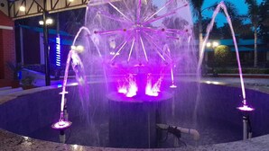 A water fountain with multiple jets and a central illuminated structure, surrounded by a paved area and palm trees. - Arunya Hotel & Resort (Rajgir)