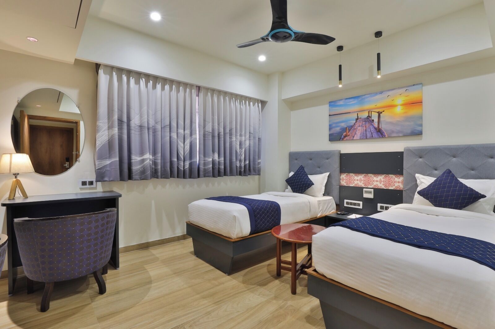 A hotel room with two beds, a desk, a chair, a ceiling fan, and a painting of a sunset.