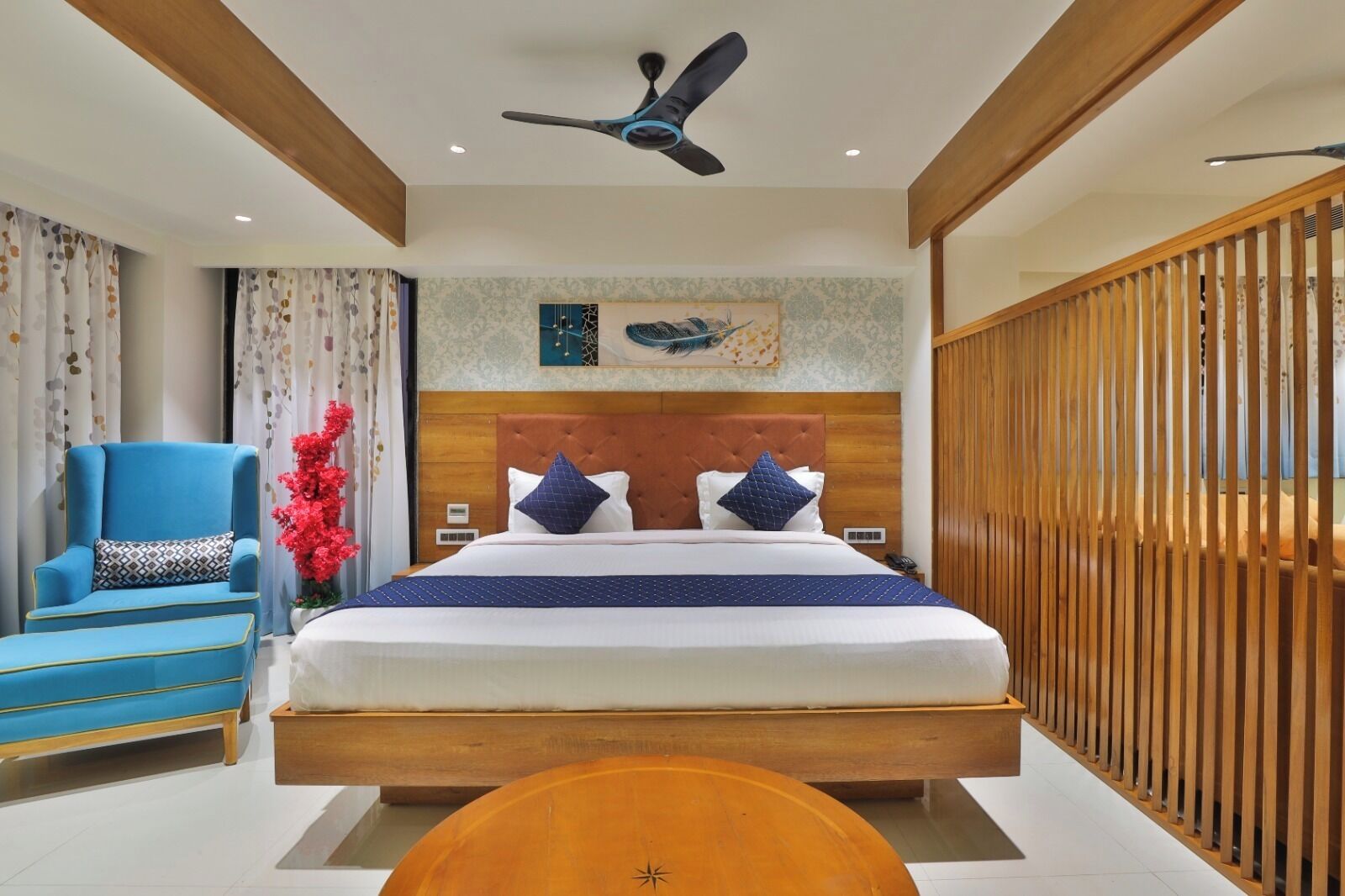 A hotel room with a bed, a chair, a wooden floor, a ceiling fan, and a decorative wall piece.