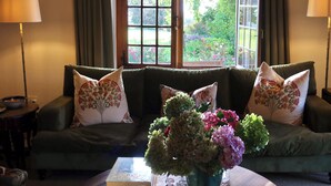 A cozy living room with a green sofa, floral cushions, a table with a book and a vase of flowers, and a window with a view of greenery. - Navarre Residence (Stellenbosch)