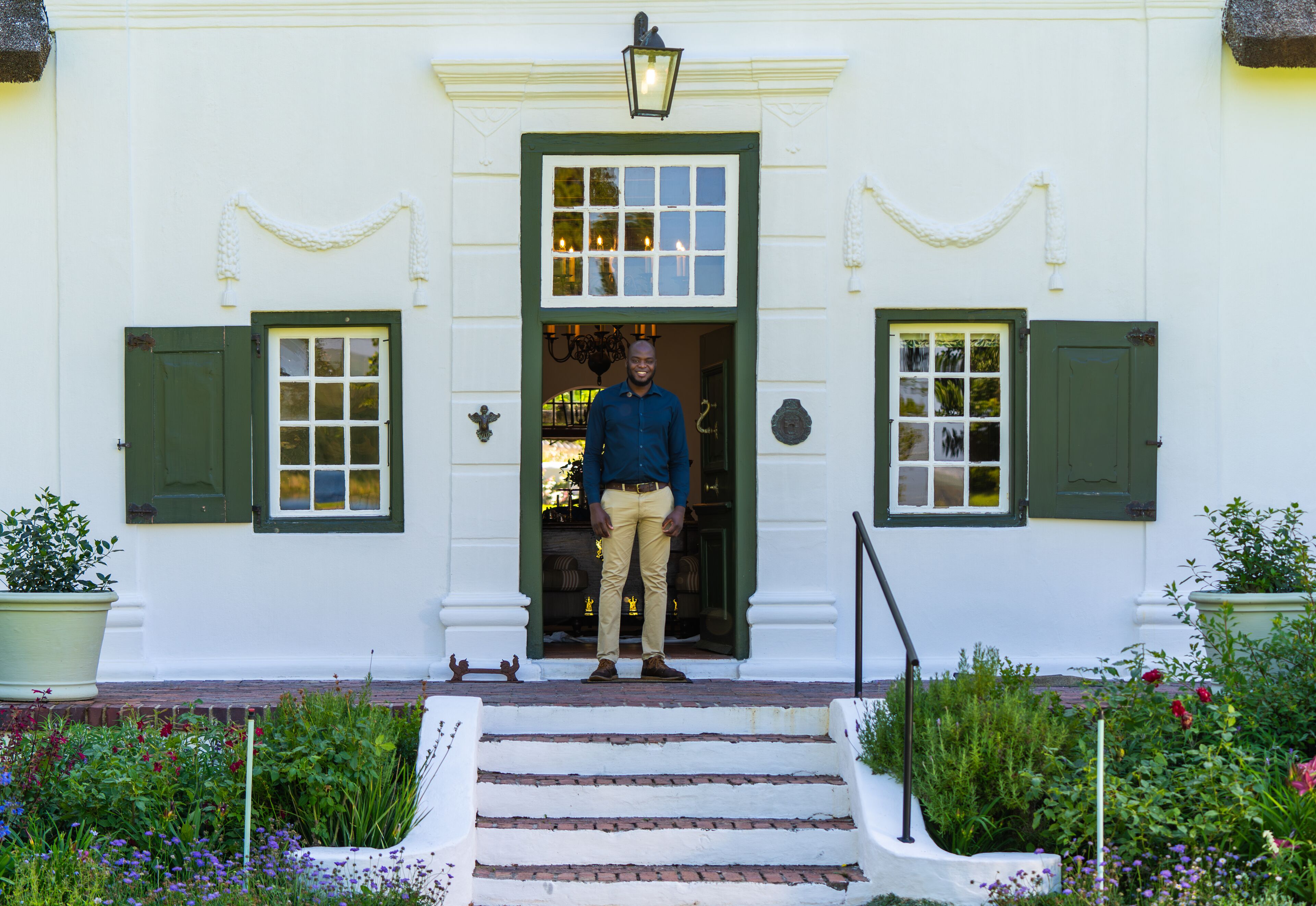 A man standing at the entrance of a white building with green shutters, a set of steps leading up to the door, and a garden with various plants and flowers.