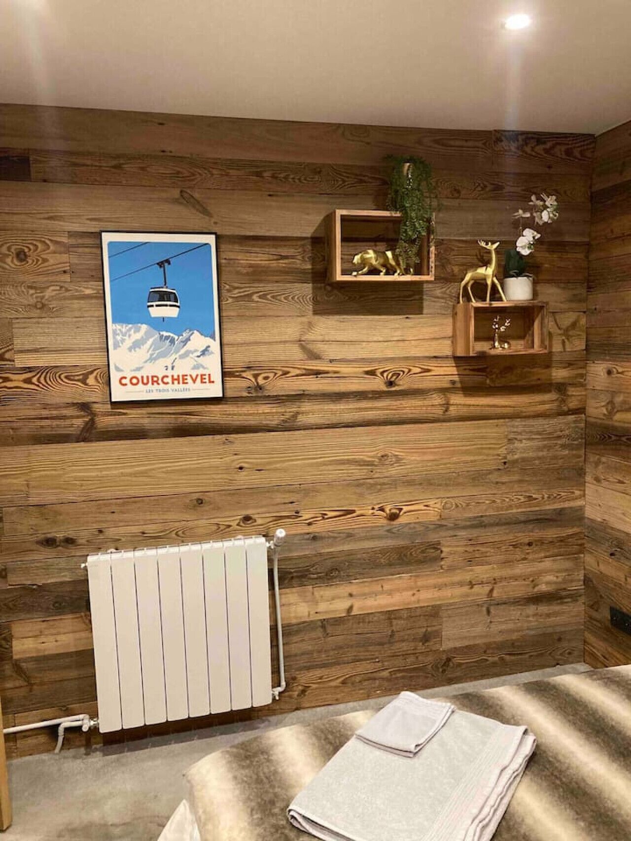 Una habitación con paneles de madera, un cartel de Courchevel, un radiador y una toalla doblada cuidadosamente sobre el suelo.