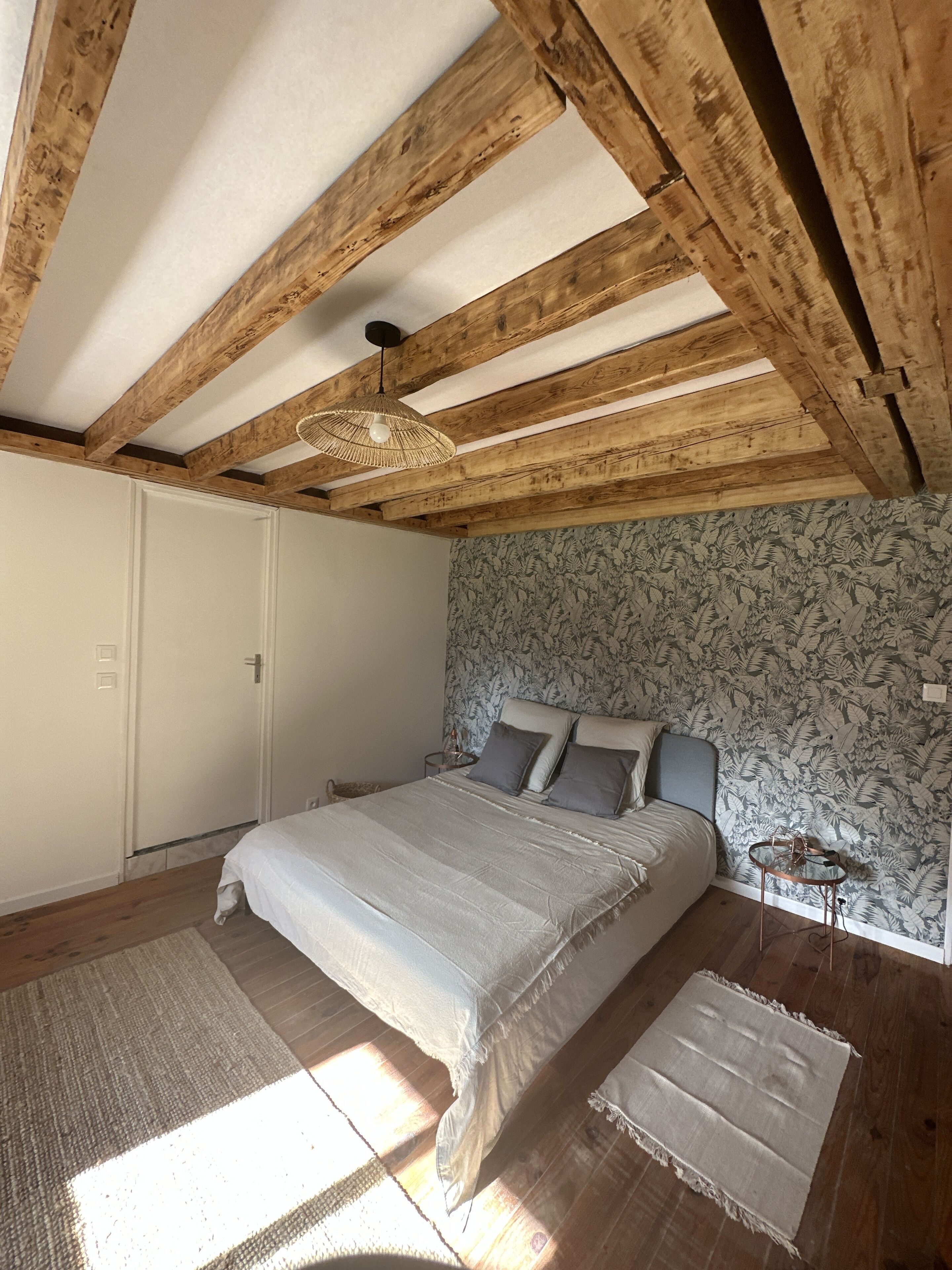 A bedroom with a wooden ceiling, a bed with a grey bedspread, a nightstand, and a patterned wallpaper.