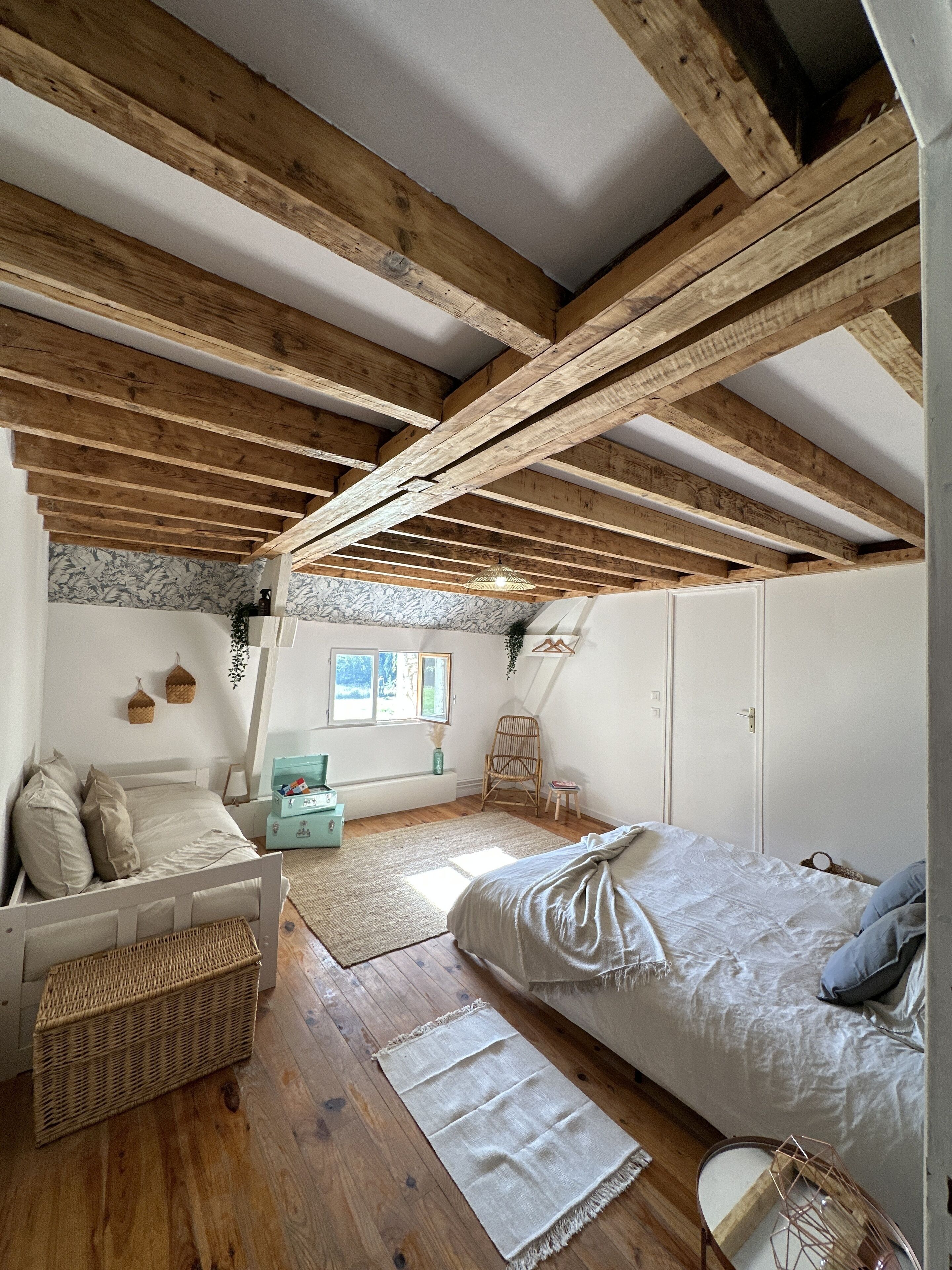 A cozy attic bedroom with exposed wooden beams, a bed, a small seating area, and a window with a view.
