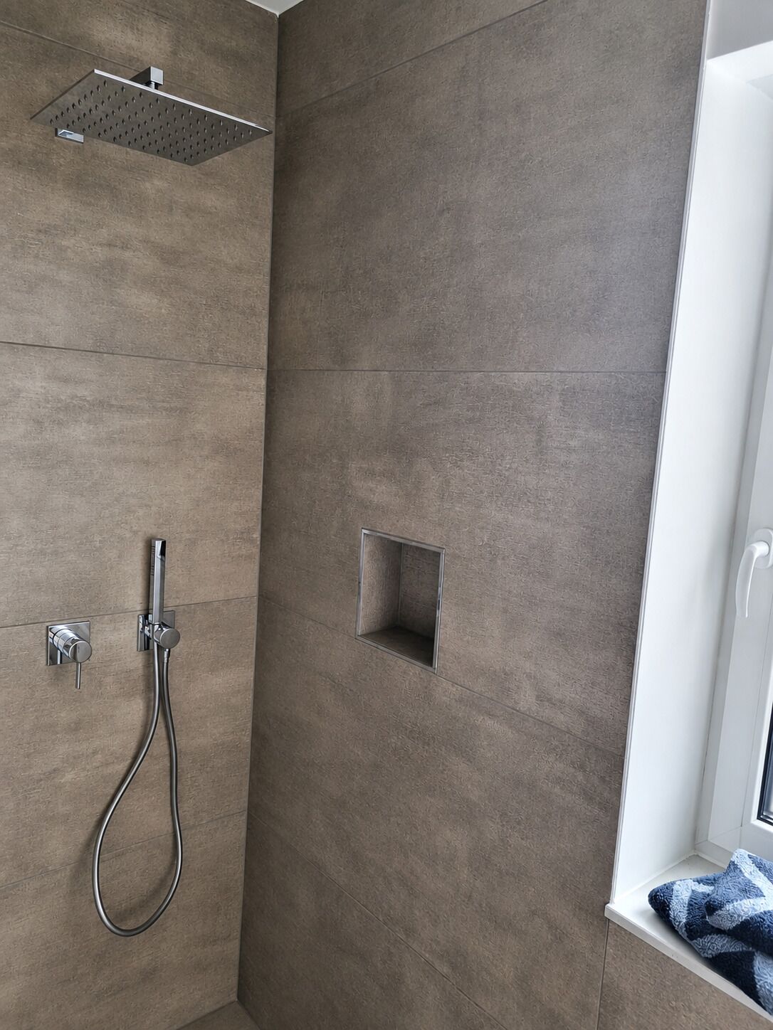 Área de duche com chuveiro manual, painel de controlo na parede e um pequeno respiradouro quadrado.