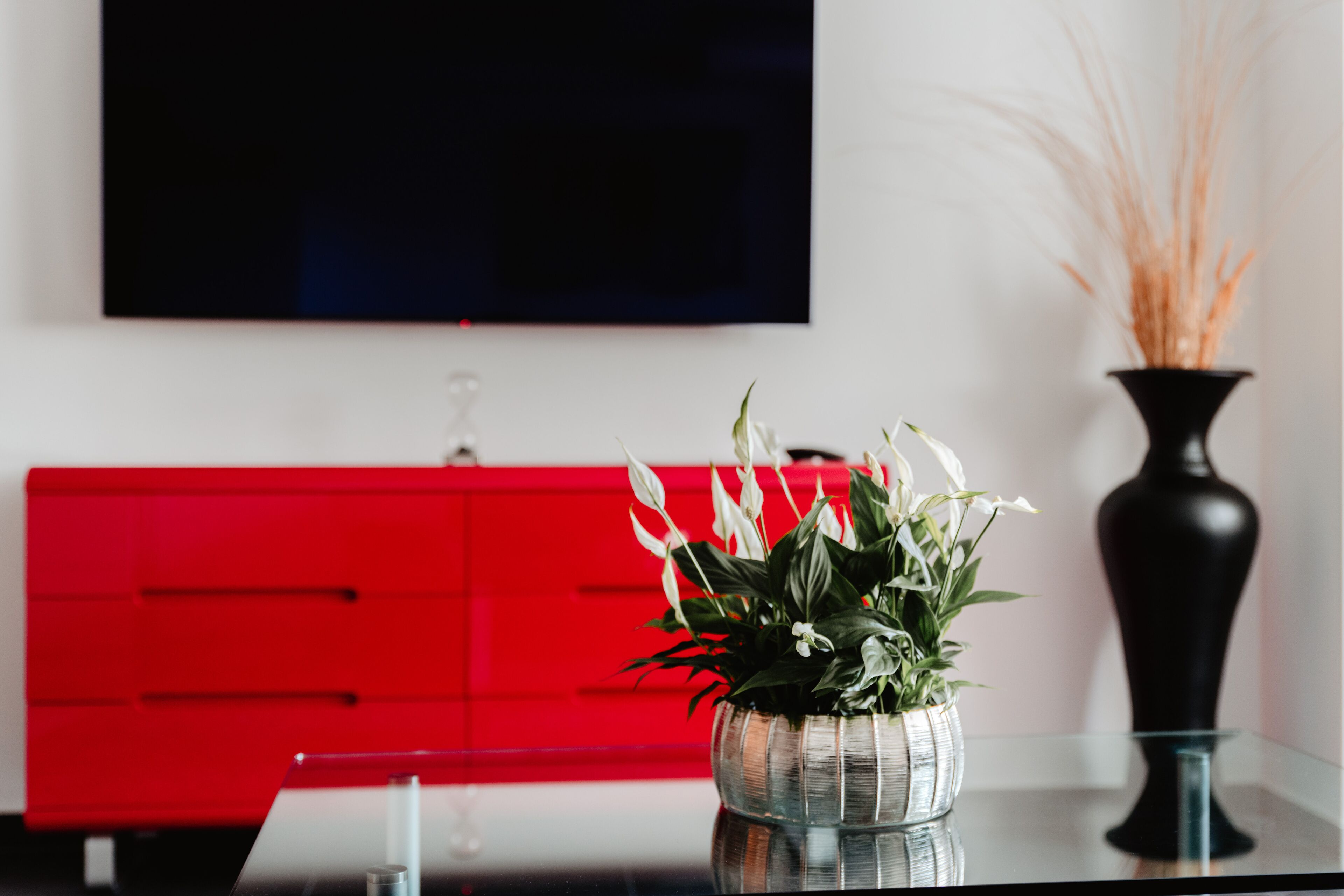 Een moderne woonkamer met een glazen salontafel, een rode kast, een zwarte vaas met gedroogde planten en een flatscreen televisie aan de muur.