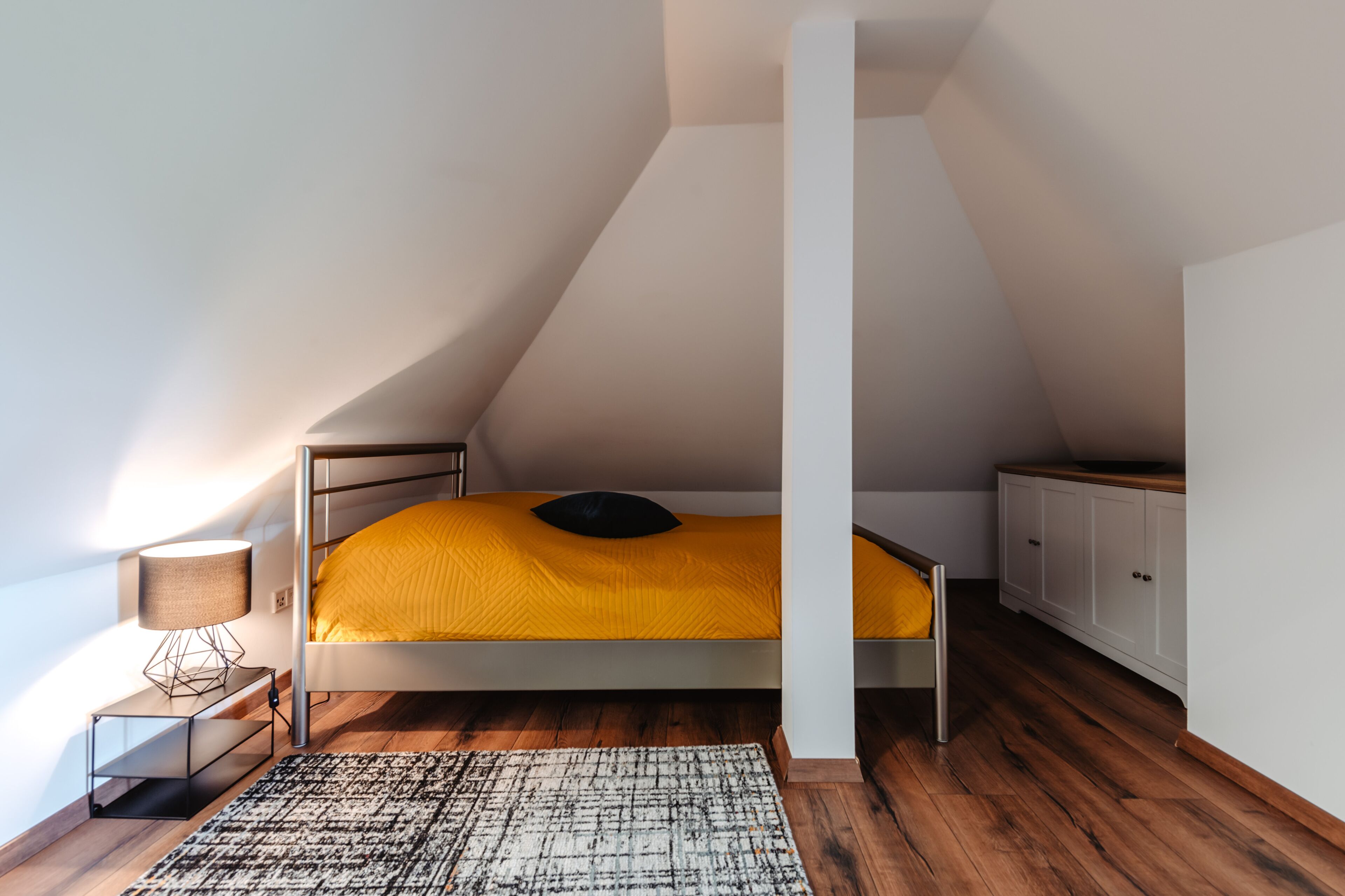 A bedroom with a bunk bed, a bedside table with a lamp, a patterned rug, wooden flooring, and a white wardrobe.