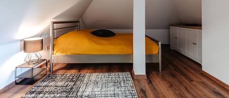A bedroom with a bunk bed, a bedside table with a lamp, a patterned rug, wooden flooring, and a white wardrobe.