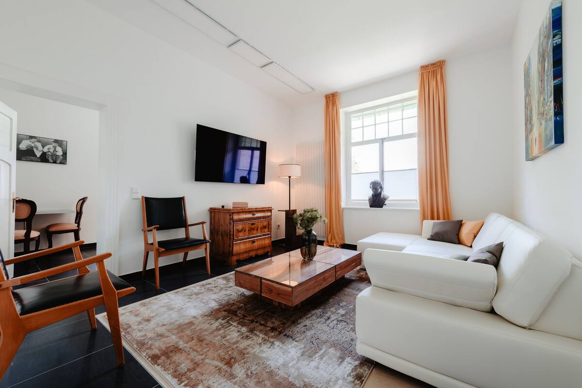 A modern living room with a white sofa, wooden coffee table, and two wooden chairs. A flat-screen TV is mounted on the wall, and there is a lamp on a side table.