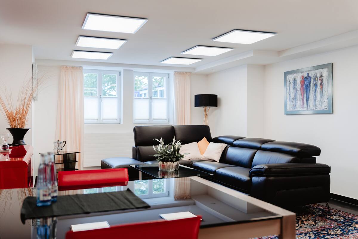 A modern living room with a black leather sofa, a glass coffee table, and a red dining table. There are large windows with curtains, a flat-screen TV mounted on the wall, and a painting above the sofa.
