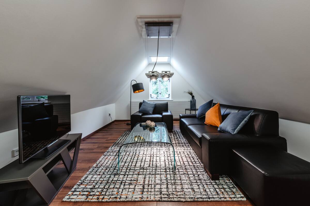 A modern loft apartment with a high ceiling, a large window, a flat-screen TV, a black leather sofa, and a patterned rug.